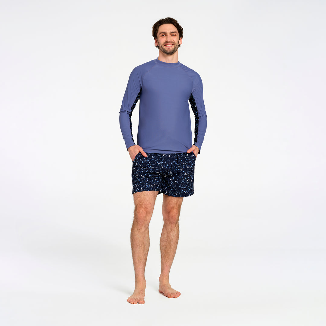 A man modeling a long-sleeve athletic rash guard in slate blue with black side panels and matching dark patterned swim shorts, standing against a plain white studio background.