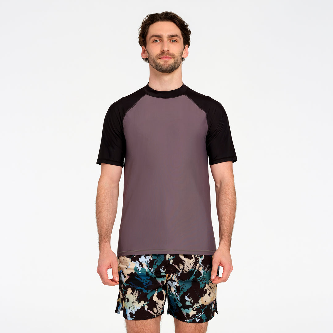 A man modeling a short-sleeve athletic rash guard with a solid dark gray body and black raglan sleeves, paired with patterned swim trunks, against a plain white studio background.