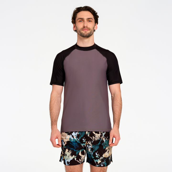 A man modeling a short-sleeve athletic rash guard with a solid dark gray body and black raglan sleeves, paired with patterned swim trunks, against a plain white studio background.