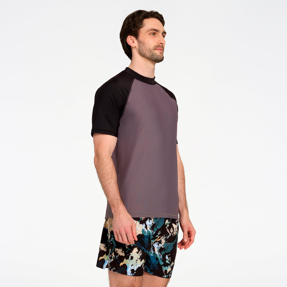 A man wearing a two-tone raglan t-shirt with a dark gray body and black sleeves, paired with patterned swim trunks, photographed against a solid white studio background.