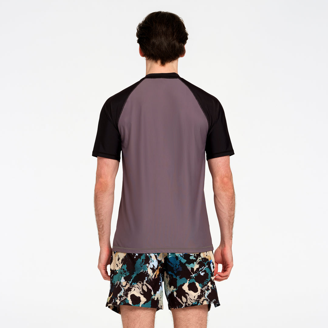 A man viewed from behind wearing a short-sleeve athletic rash guard with a muted gray-purple body and black raglan sleeves, paired with patterned swim shorts against a plain white studio background.