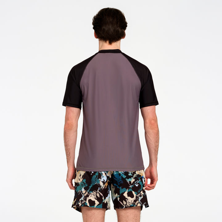 A man viewed from behind wearing a short-sleeve athletic rash guard with a muted gray-purple body and black raglan sleeves, paired with patterned swim shorts against a plain white studio background.