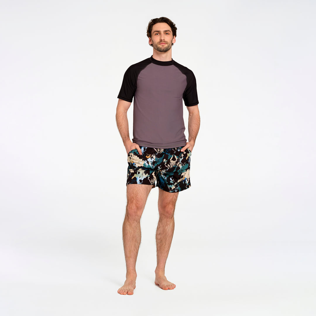 A man modeling a short-sleeve raglan t-shirt with a muted purple body and black sleeves, paired with black swim trunks featuring a multicolor abstract pattern, against a plain white studio background.