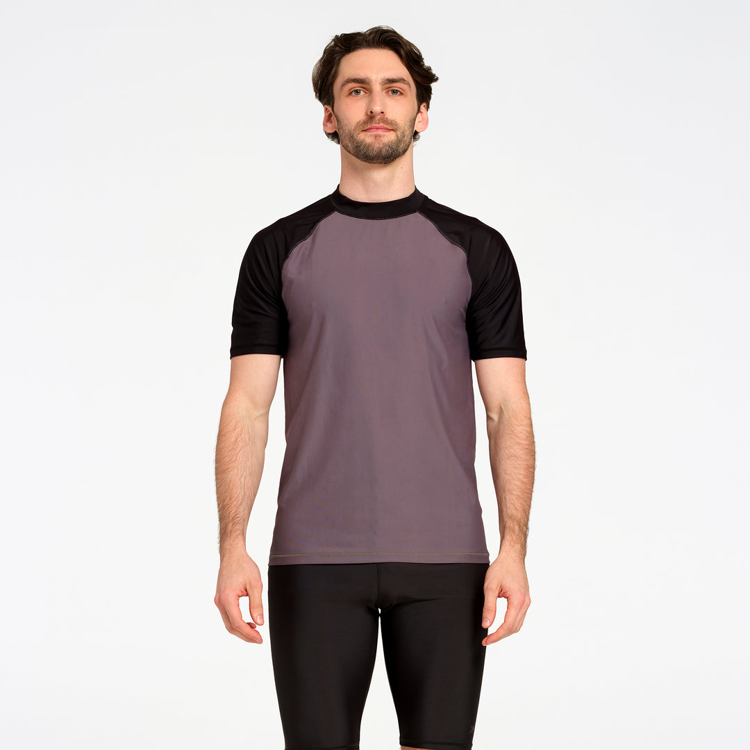 A man wearing a short-sleeve athletic shirt with a muted purple-gray body and black raglan sleeves, paired with black shorts, against a plain light gray studio background.