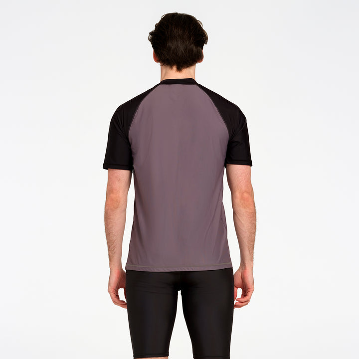 A man viewed from the back wearing a short-sleeve athletic jersey with a muted mauve-gray body and black raglan sleeves, paired with black shorts, against a plain white studio background.