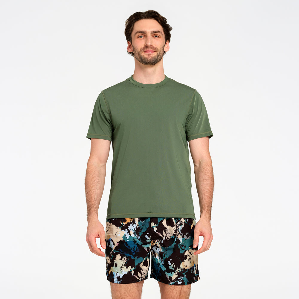 A man modeling a short-sleeve crewneck swim t-shirt in olive green, paired with patterned swim shorts, against a plain white studio background.