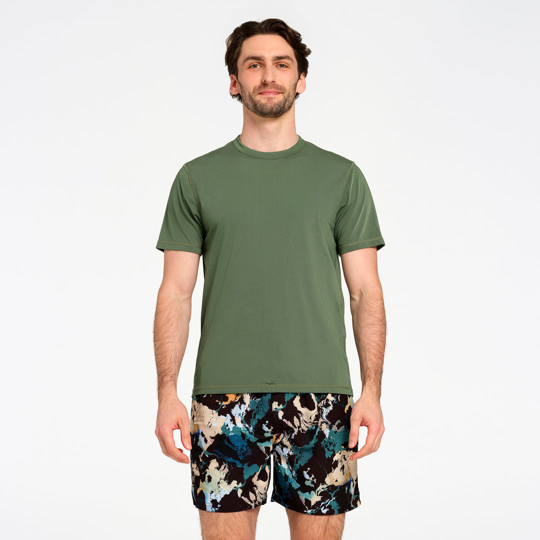 A man modeling a short-sleeve crewneck swim t-shirt in olive green, paired with patterned swim shorts, against a plain white studio background.