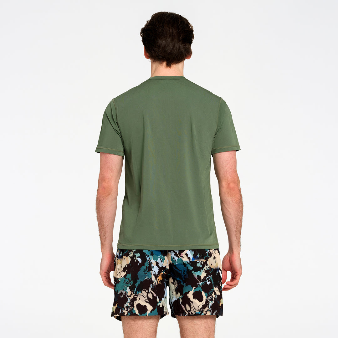 A man viewed from the back wearing an olive green short-sleeve swim t-shirt and black swim boardshorts with a blue, beige, and white abstract pattern