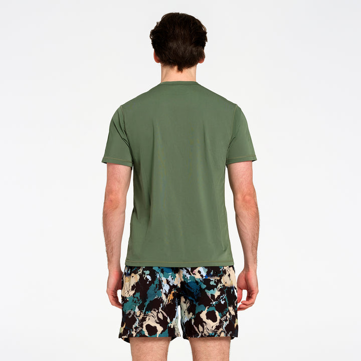 A man viewed from the back wearing an olive green short-sleeve swim t-shirt and black swim boardshorts with a blue, beige, and white abstract pattern