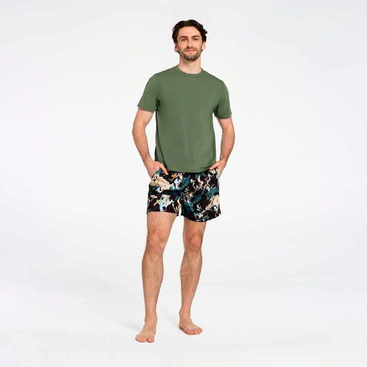 A man standing against a white studio background wearing an olive green short-sleeve swim t-shirt and black swim shorts with a colorful abstract pattern.