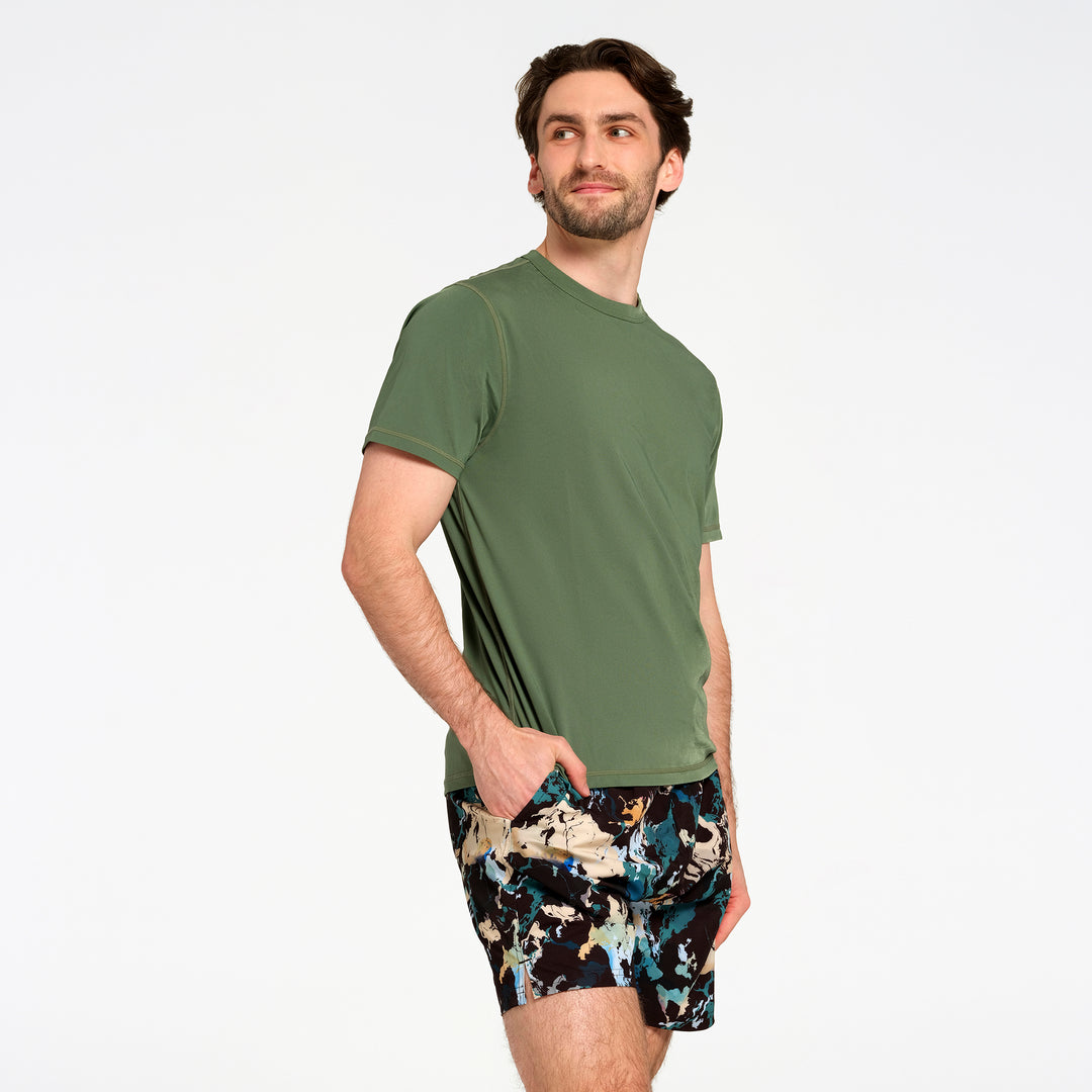 A man wearing an olive green short-sleeve swim t-shirt and black swim trunks with a blue, beige, and white abstract pattern, standing against a plain white studio background.