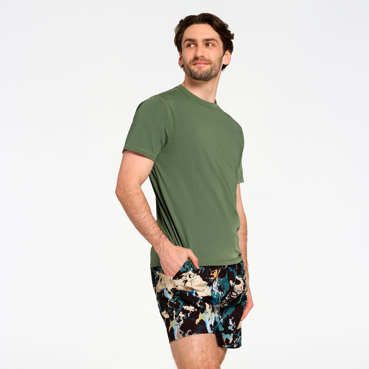 A man wearing an olive green short-sleeve swim t-shirt and black swim trunks with a blue, beige, and white abstract pattern, standing against a plain white studio background.