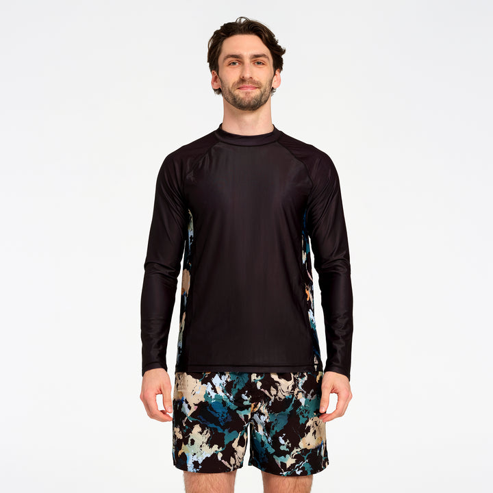 A man modeling a black long-sleeve rash guard with abstract blue, beige, and black patterned side panels, paired with matching patterned swim shorts, against a plain white studio background.