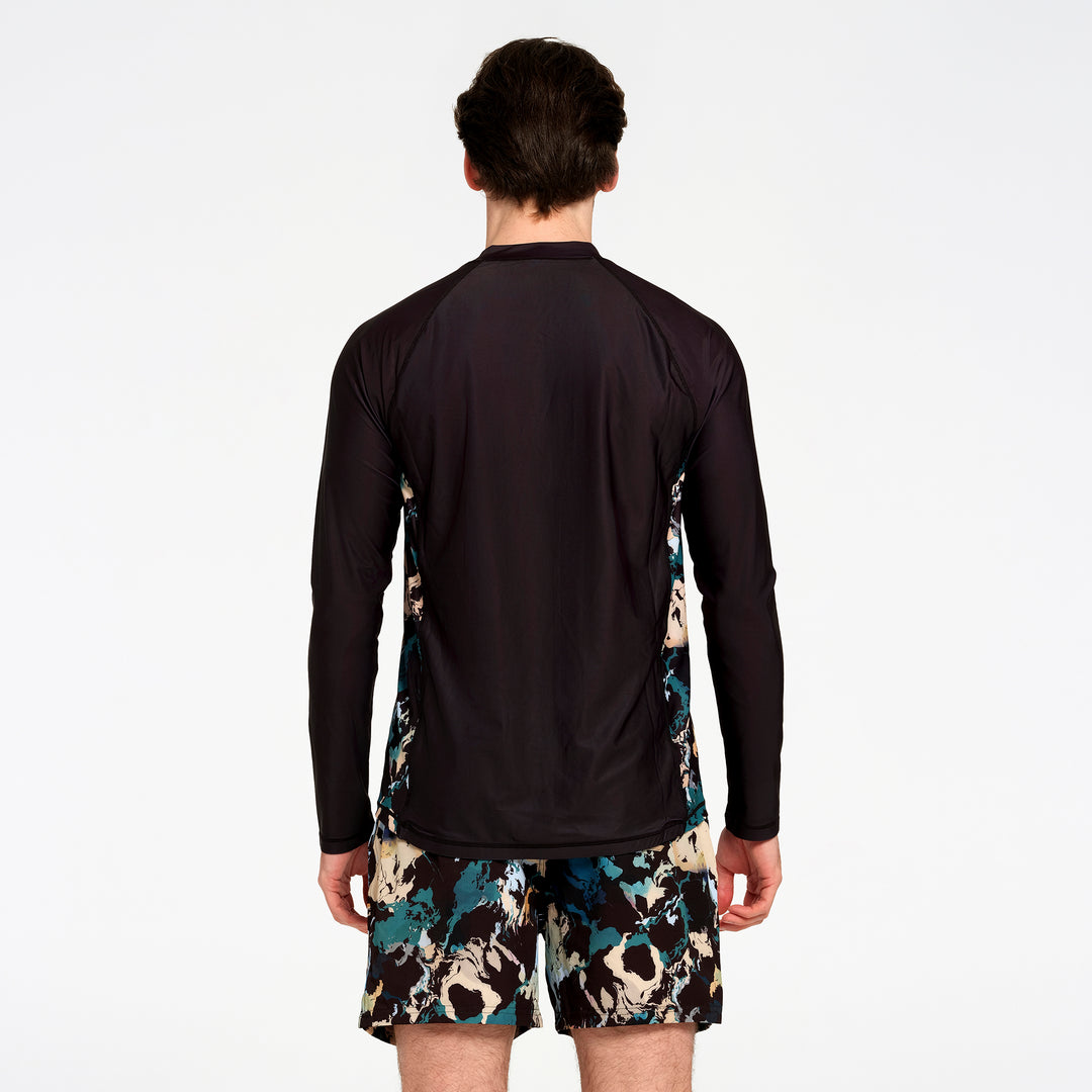 A man viewed from the back wearing a black long-sleeve athletic top with teal and beige abstract patterned side panels, paired with matching patterned shorts, against a plain white studio background.