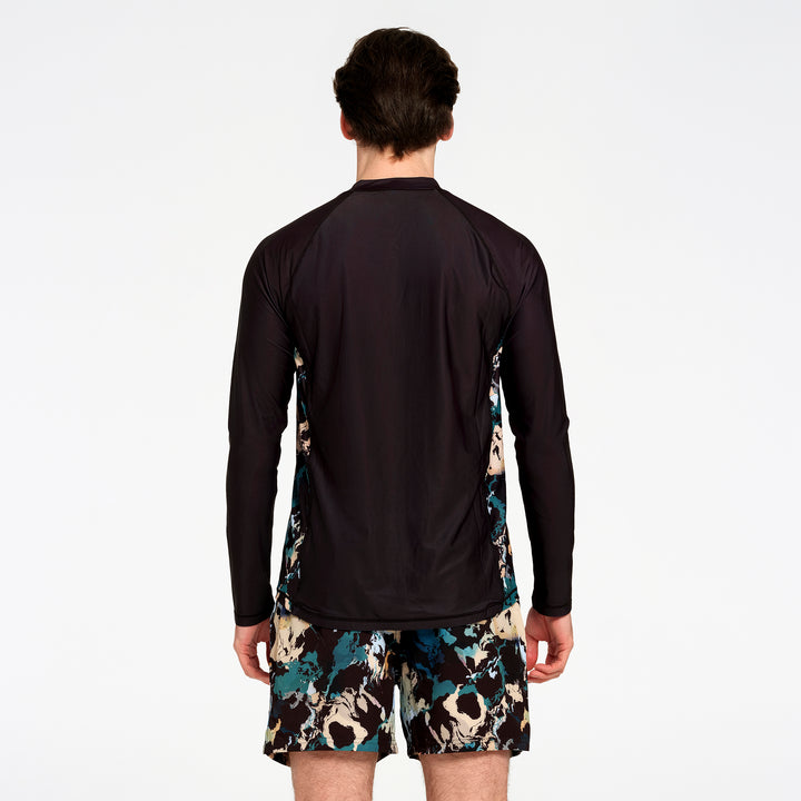 A man viewed from the back wearing a black long-sleeve athletic top with teal and beige abstract patterned side panels, paired with matching patterned shorts, against a plain white studio background.