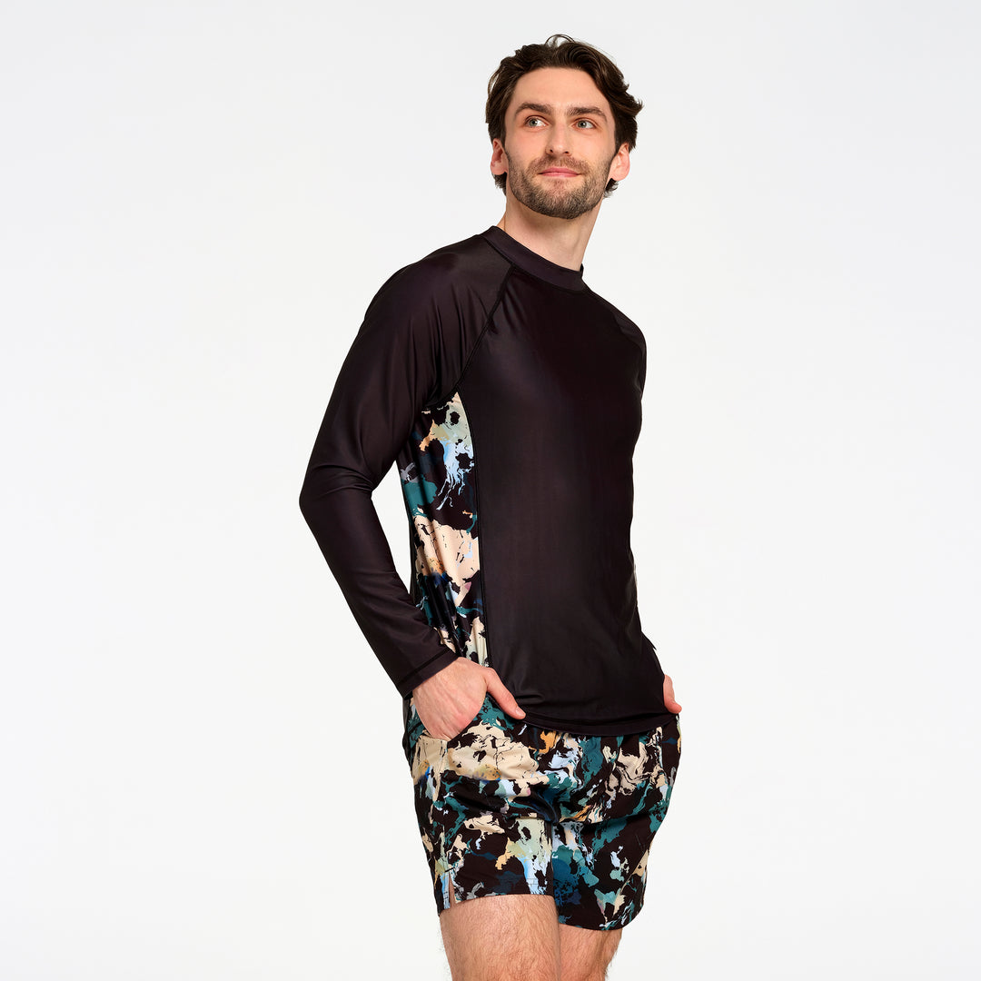 A man modeling a long-sleeve swim rash guard and matching swim shorts against a plain white studio background. The rash guard is black with a multicolor abstract pattern on the sides, and the swim shorts feature a matching multicolor abstract print.