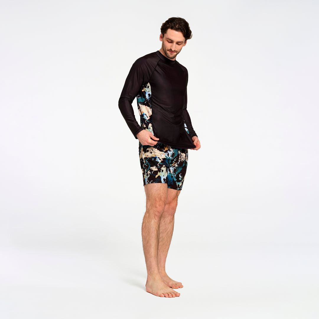 A man modeling a black long-sleeve swim rash guard with abstract blue, beige, and black patterned shorts against a plain white studio background.