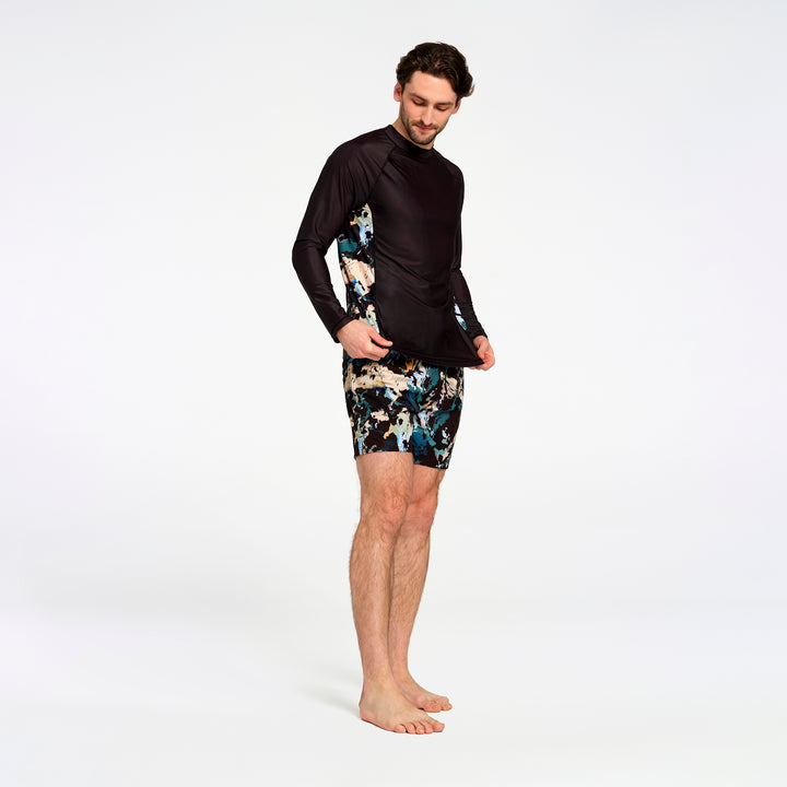 A man modeling a black long-sleeve swim rash guard with abstract blue, beige, and black patterned shorts against a plain white studio background.
