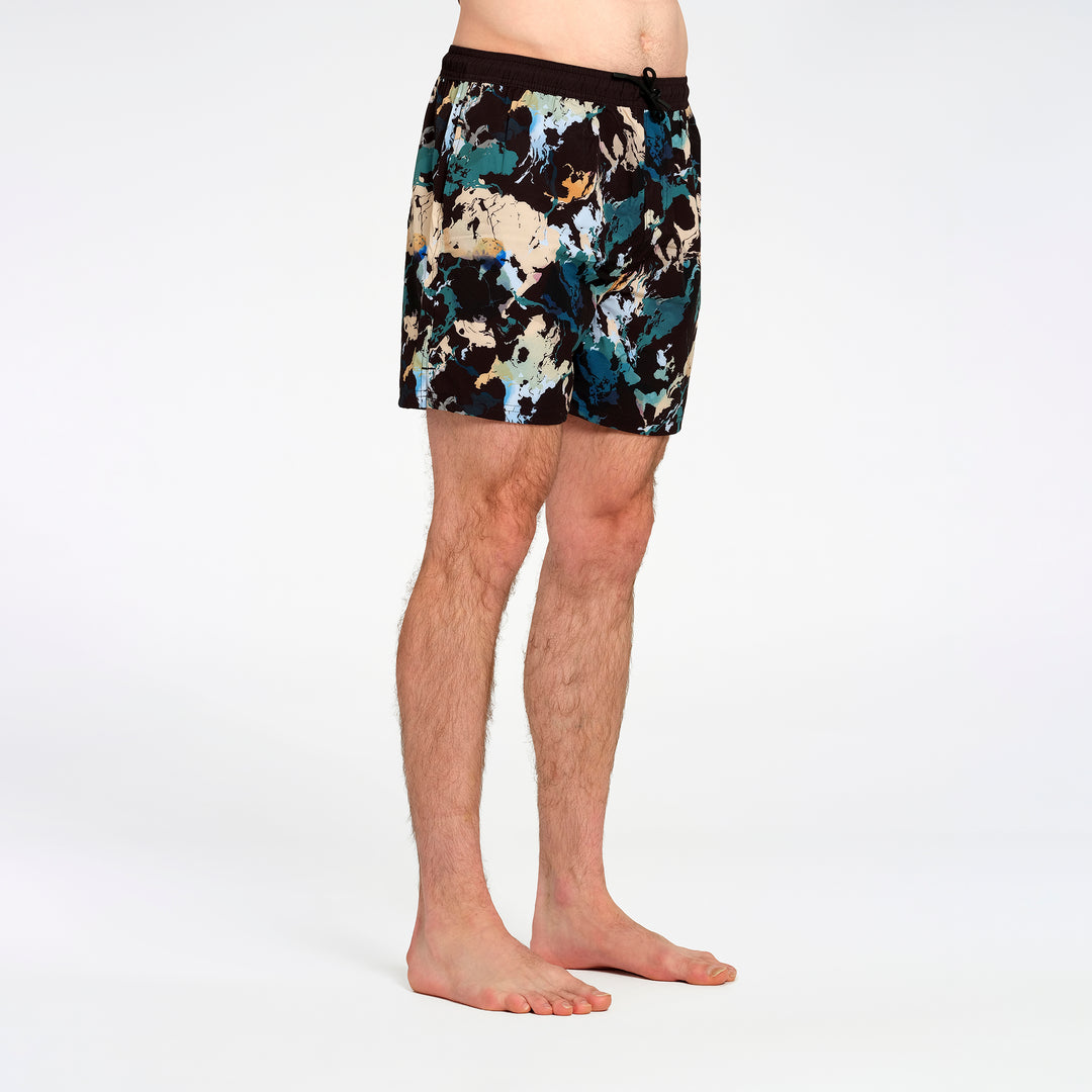 Men's swim shorts with an abstract multicolor print in teal, beige, and black, modeled on a person against a white studio background.