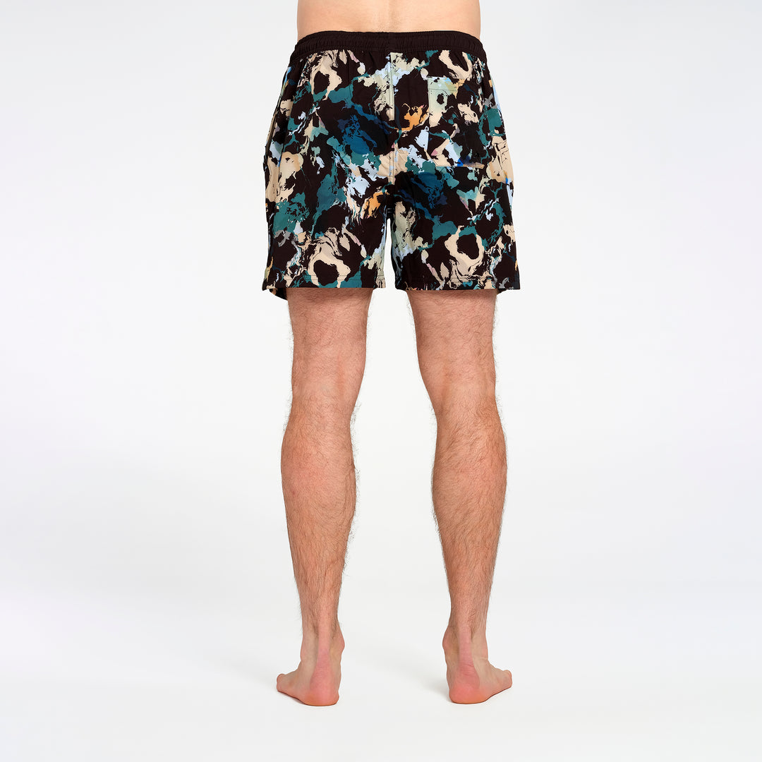 Rear view of a man wearing patterned swim shorts with a black, teal, beige, and white abstract print on a white studio background.