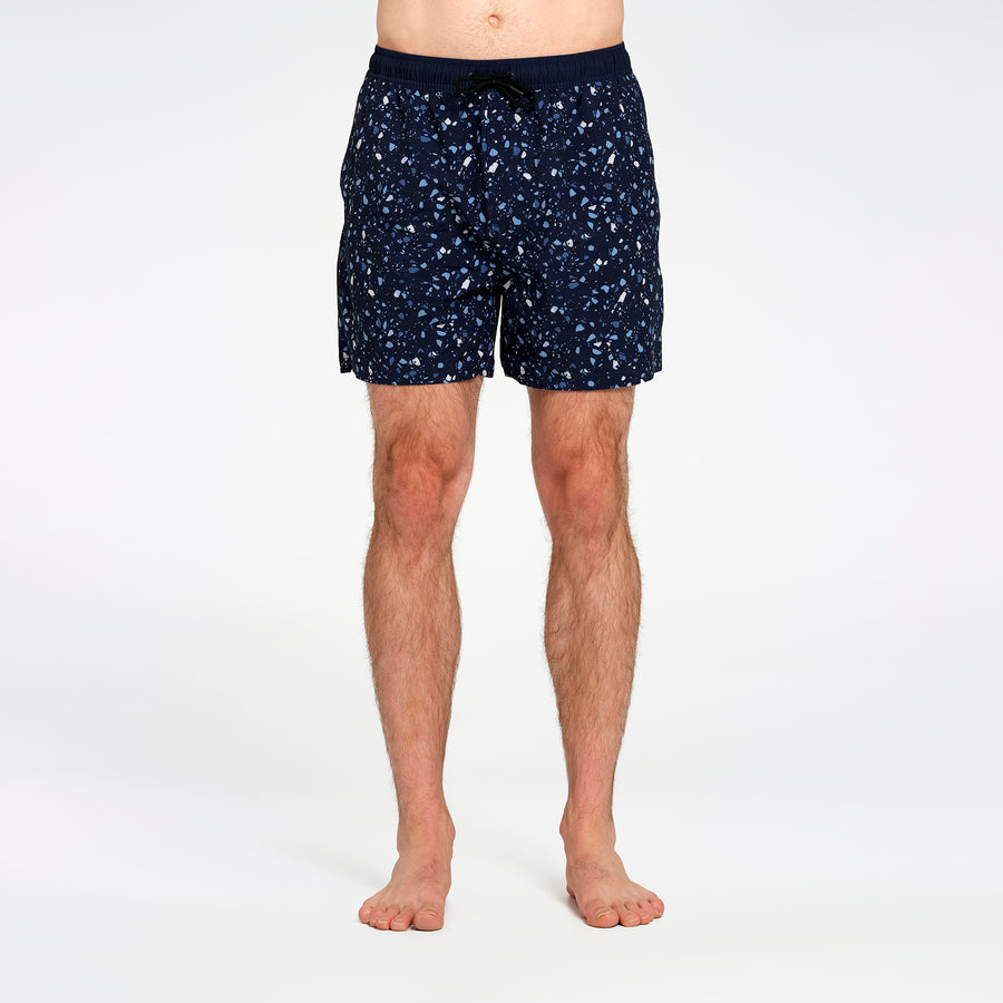 A man's legs and lower torso are shown wearing dark navy blue swim shorts with a small white speckled pattern, standing against a plain white studio background.