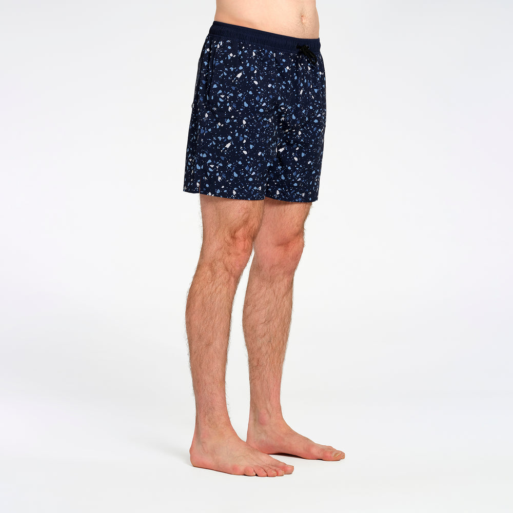 A side profile view of a man wearing navy blue swim shorts with a white and light blue speckled pattern, standing against a plain white studio background.