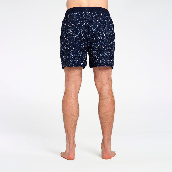 Rear view of a person wearing dark navy patterned shorts with small white and light blue speckles, shown against a plain light gray studio background.
