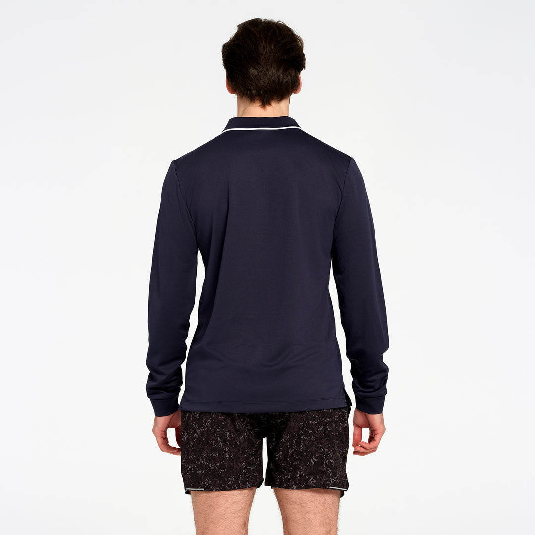 A man viewed from the back wearing a navy blue long-sleeve polo shirt with white piping on the collar, paired with black patterned shorts, against a plain white background.