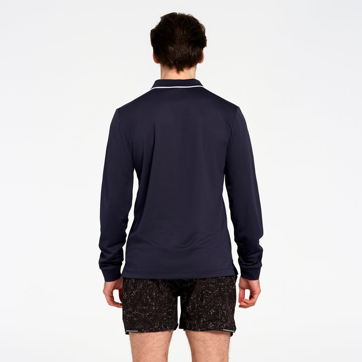 A man viewed from the back wearing a navy blue long-sleeve polo shirt with white piping on the collar, paired with black patterned shorts, against a plain white background.