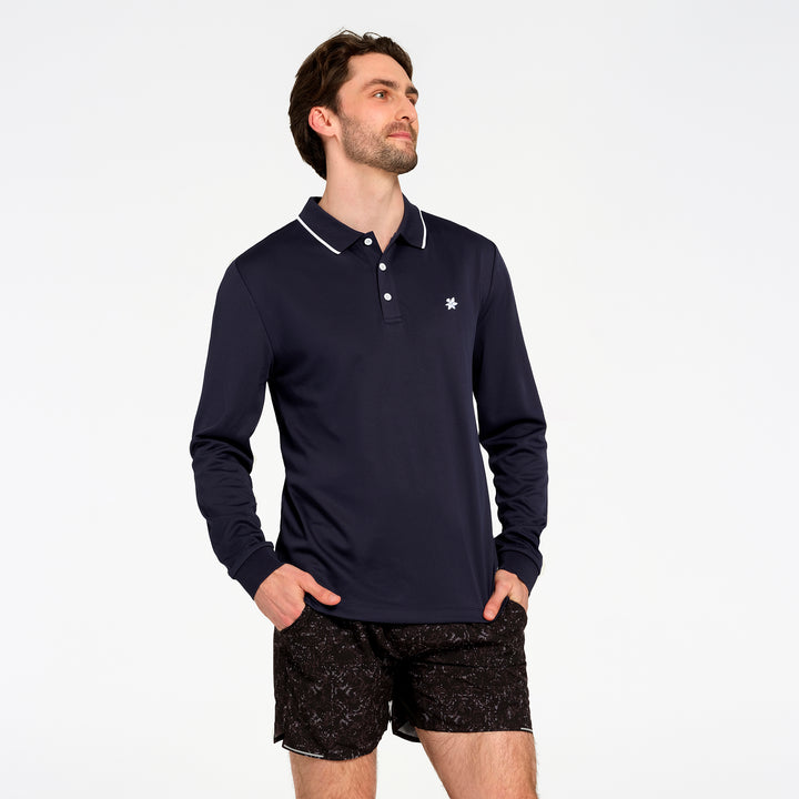 A man wearing a navy blue long-sleeve polo shirt with white piping on the collar and a small white embroidered logo on the chest, paired with black patterned shorts, against a plain white background.
