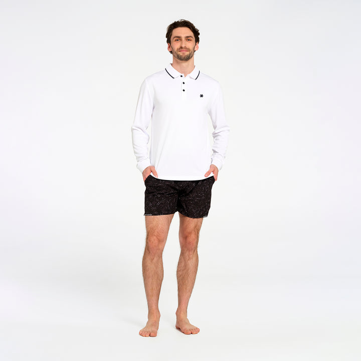 A man modeling a white long-sleeve polo shirt with black trim and a small black chest logo, paired with black patterned shorts, standing against a plain white studio background.