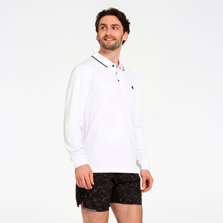 A man wearing a white long-sleeve polo shirt with a black collar trim and black patterned shorts, standing against a plain white studio background.