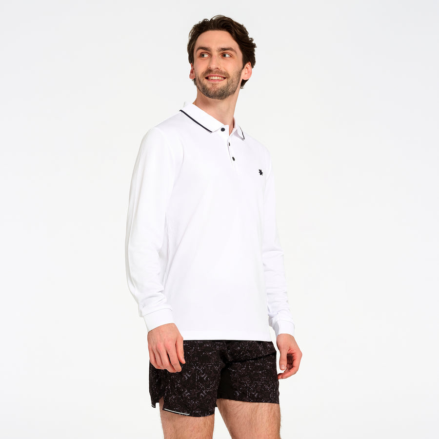 A man wearing a white long-sleeve polo shirt with a black collar trim and black patterned shorts, standing against a plain white studio background.