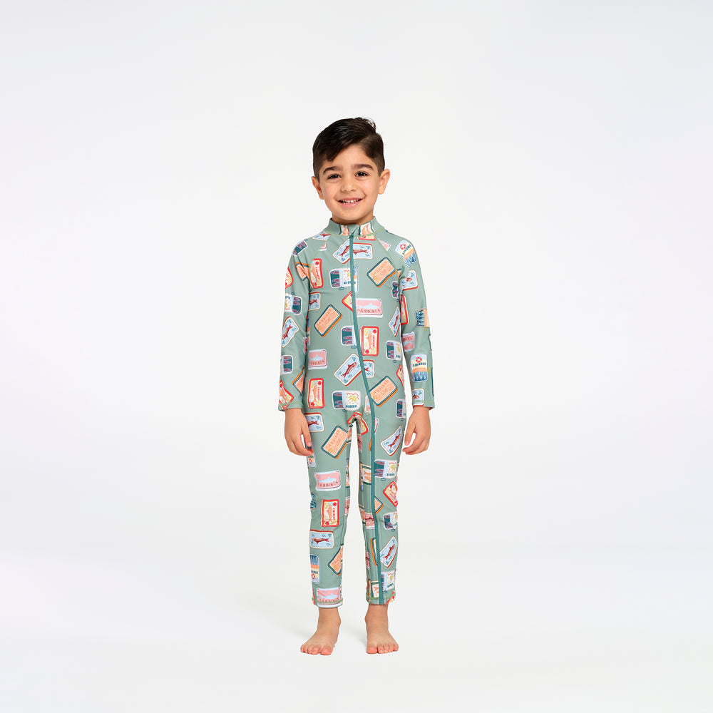A young boy stands barefoot against a plain white studio background, wearing a long-sleeve, high-neck swimsuit with an all-over retro sardine print in sage green, pink, and orange.
