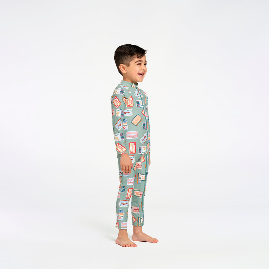 A young boy stands in profile wearing a full-length, long-sleeve swim suit in a sage green color with a colorful retro sardine print, against a plain white studio background.