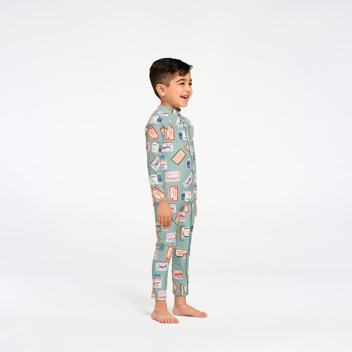 A young boy stands in profile wearing a full-length, long-sleeve swim suit in a sage green color with a colorful retro sardine print, against a plain white studio background.
