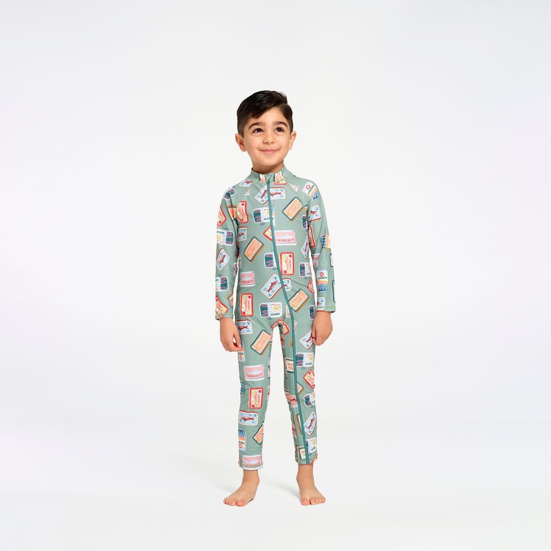 A young boy stands barefoot against a plain white studio background, wearing a long-sleeve, full-length one-piece swimsuit with an allover retro sardine print on a muted sage green base.