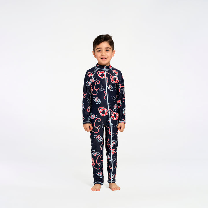 A young boy stands barefoot against a plain white studio background, wearing a long-sleeve zip-front one-piece swimsuit with a nautical print. The fabric is dark navy with red, red, and white illustrations of boats, life buoys, and waves.