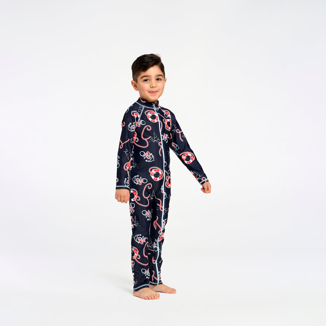 A young boy stands barefoot against a plain white studio background, wearing a long-sleeve zip-front swimsuit in dark navy with a repeating nautical print in red, white, and light blue.