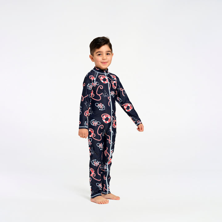 A young boy stands barefoot against a plain white studio background, wearing a long-sleeve zip-front swimsuit in dark navy with a repeating nautical print in red, white, and light blue.