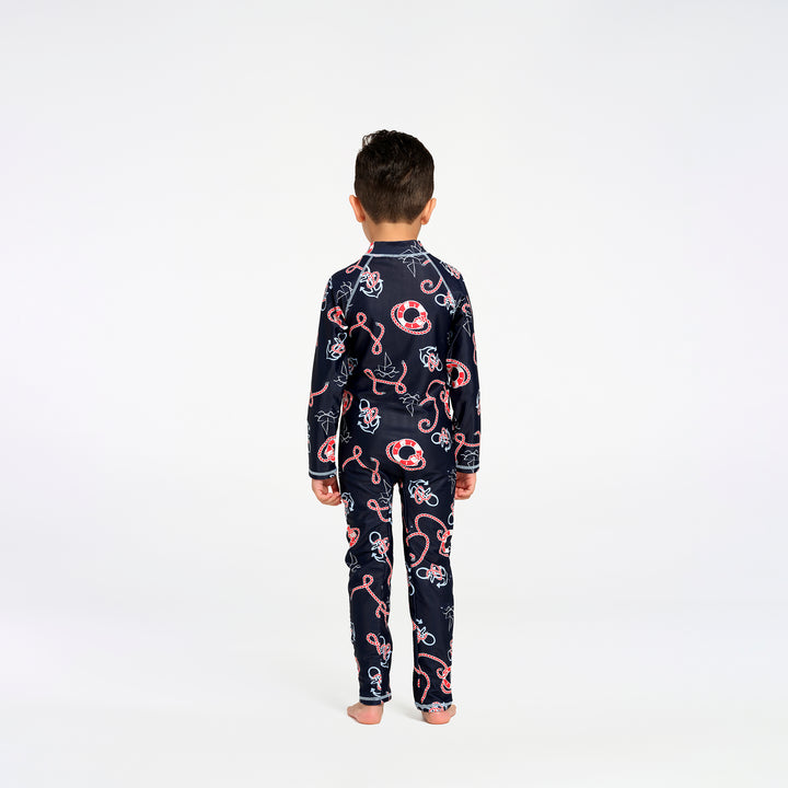 A child viewed from behind wearing a navy long-sleeve swimsuit with a red, white, and light blue nautical pattern, standing against a plain white studio background.