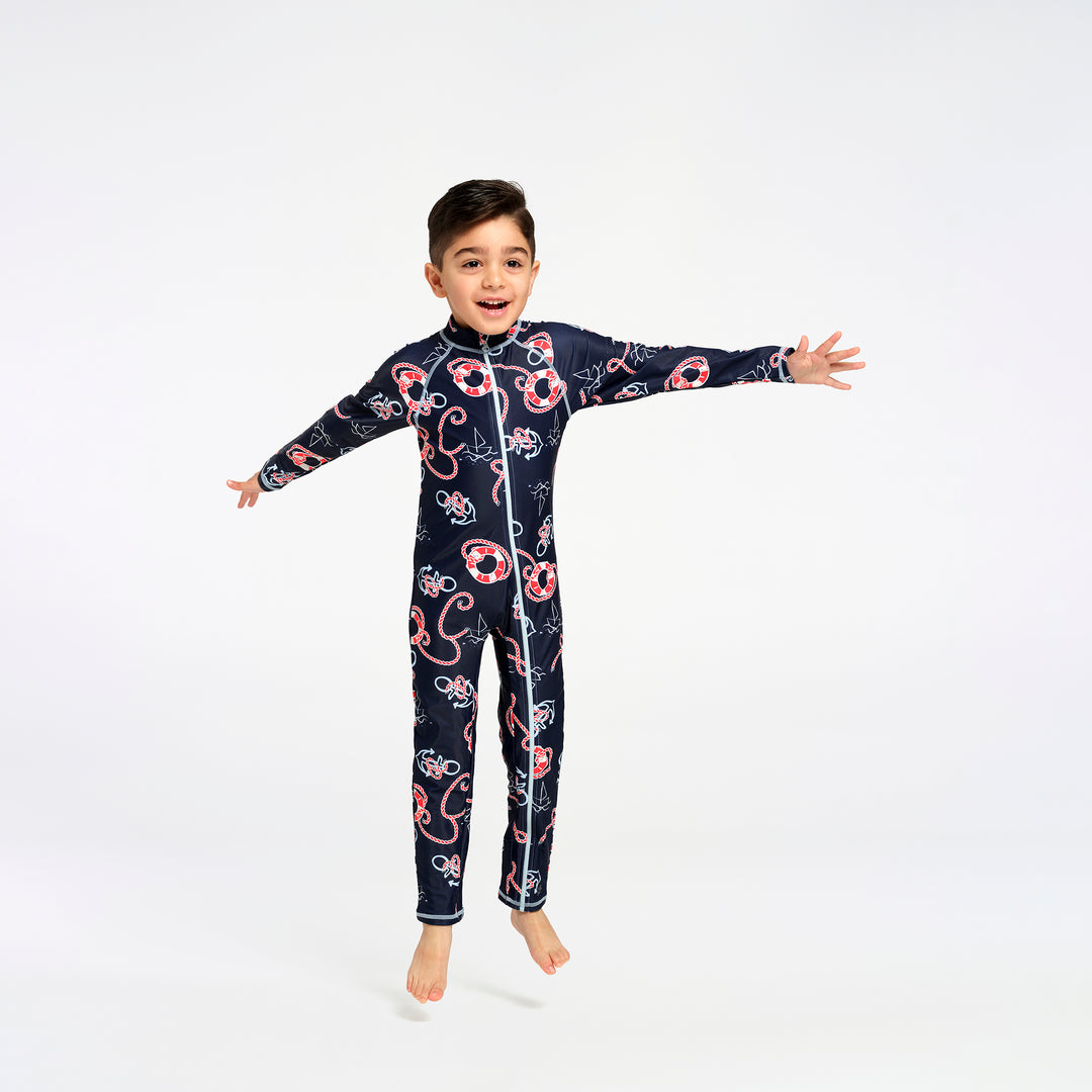 A young boy wearing a navy blue full-body one-piece swimsuit with a red and white nautical print, captured mid-jump against a seamless white studio background.