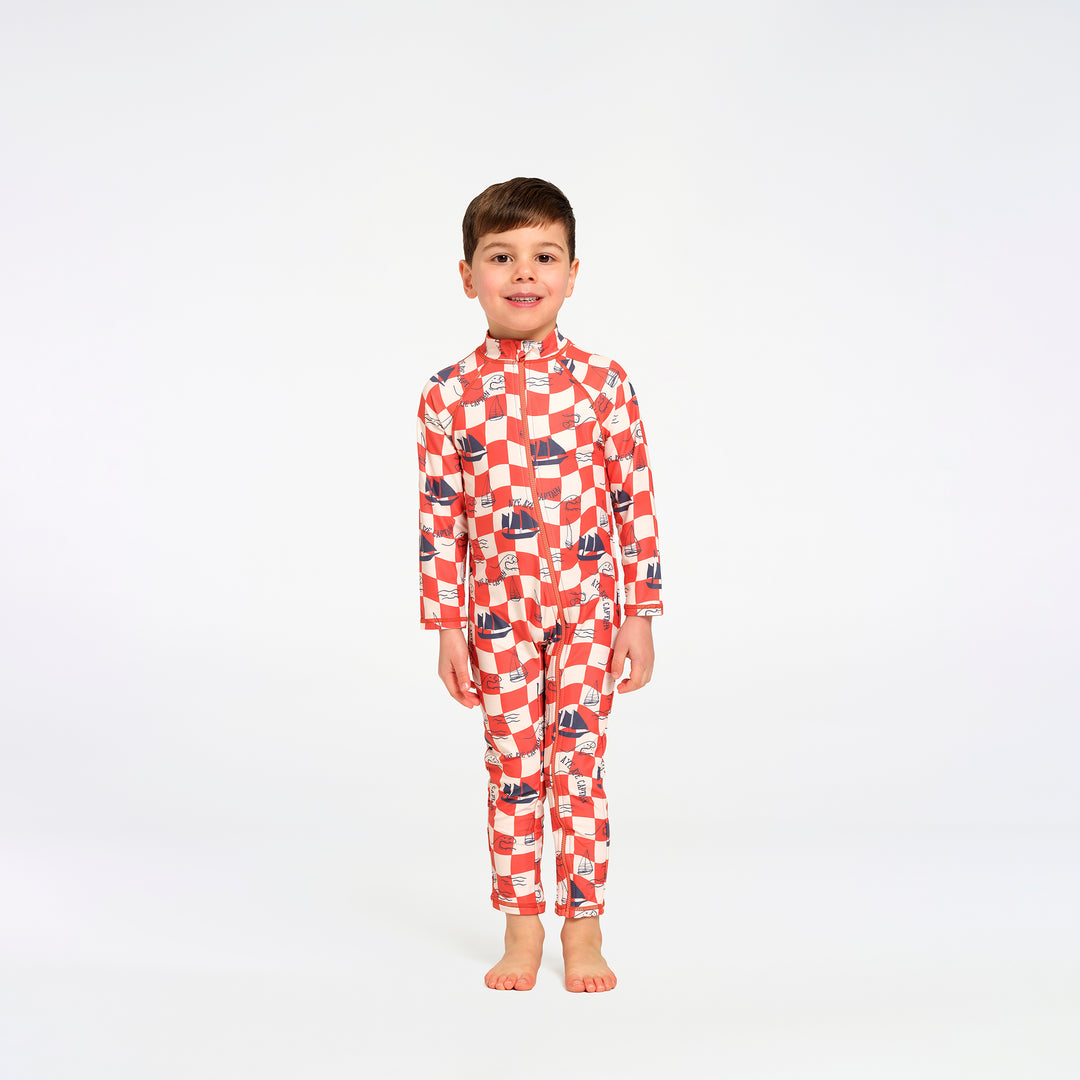 A young boy stands barefoot against a plain white studio background, wearing a long-sleeve high-neck one-piece swimsuit with a red and white checkered pattern and small blue sailboat motifs.