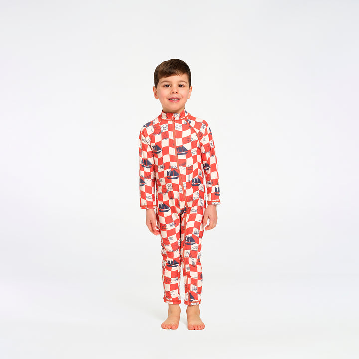 A young boy stands barefoot against a plain white studio background, wearing a long-sleeve high-neck one-piece swimsuit with a red and white checkered pattern and small blue sailboat motifs.