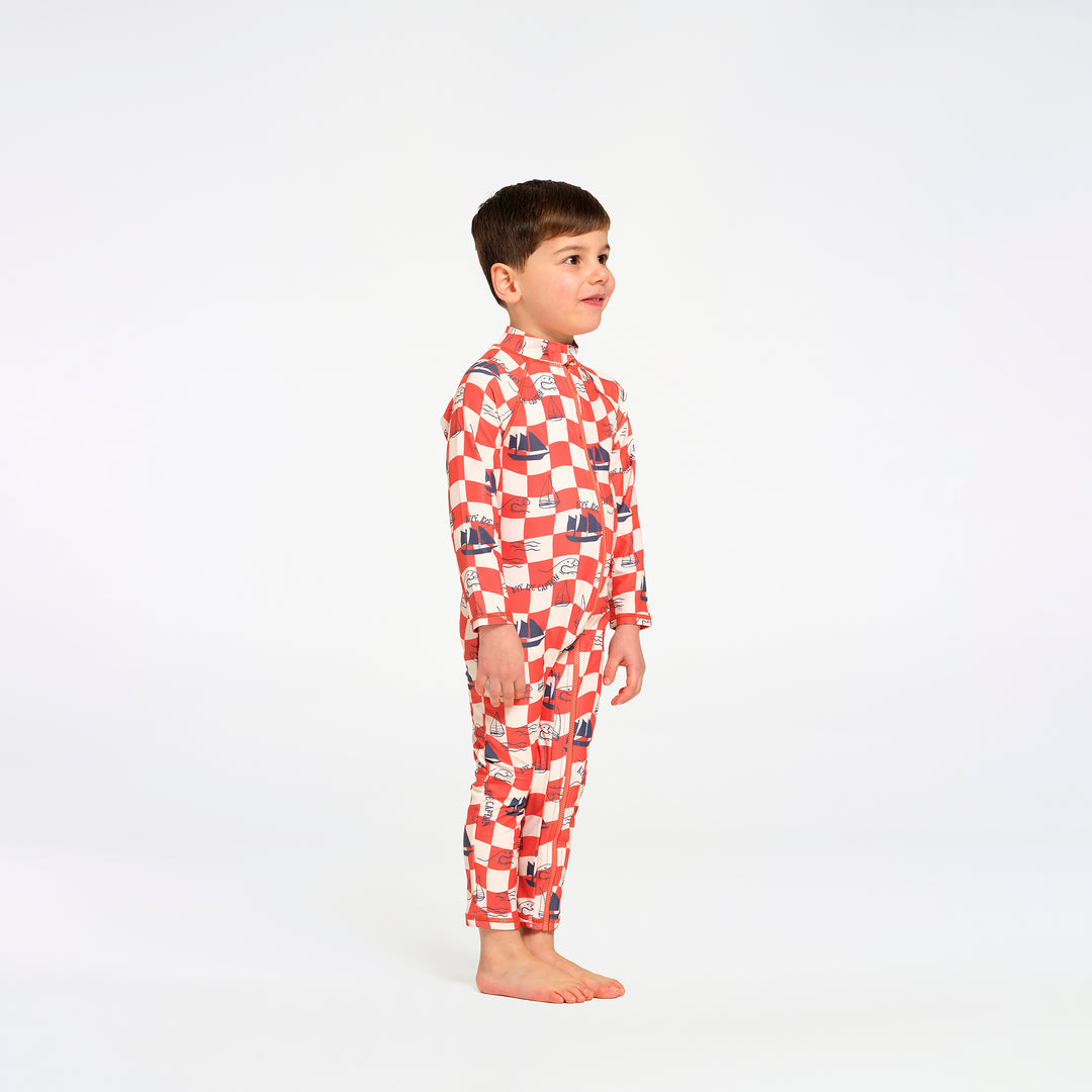 A young boy standing in profile wearing a red and white checkered long-sleeve swimsuit with small blue and red boat illustrations, photographed against a seamless white studio background.
