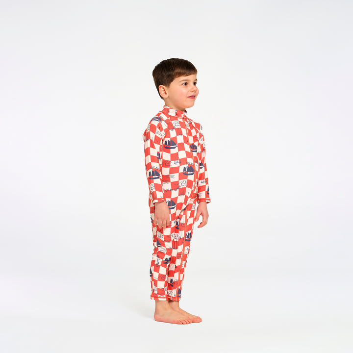 A young boy standing in profile wearing a red and white checkered long-sleeve swimsuit with small blue and red boat illustrations, photographed against a seamless white studio background.