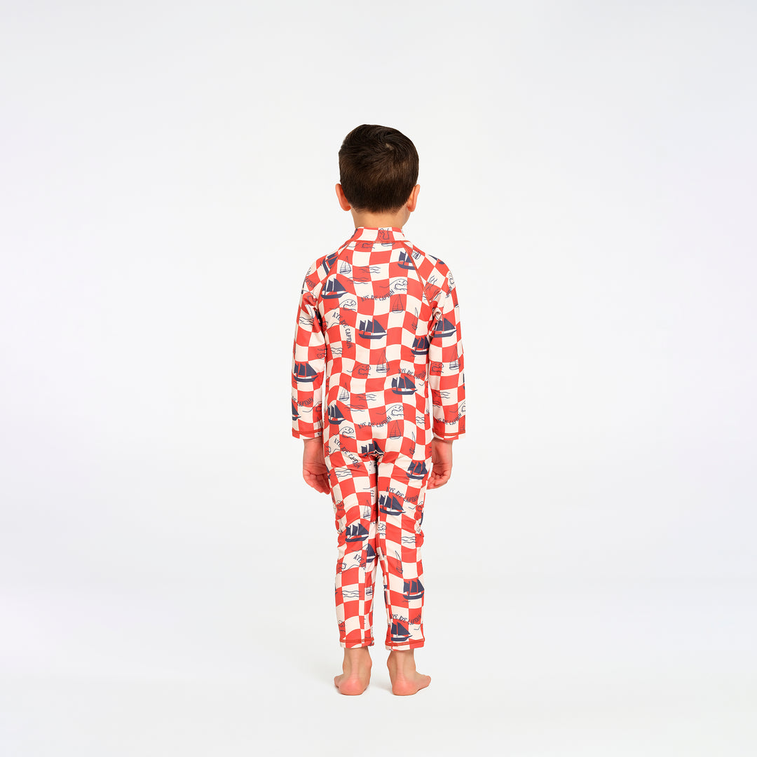 A child standing from the back, wearing a red and white checkered long-sleeve swimsuit with a small blue boat print, on a white studio background.