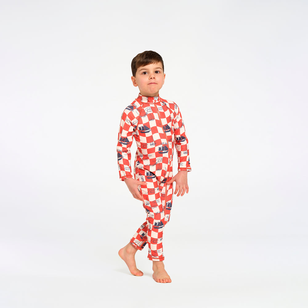 A young boy stands barefoot against a plain white studio background, wearing a long-sleeve, full-length one-piece swimsuit with an allover red and white checkered pattern and small dark blue boat motifs. 