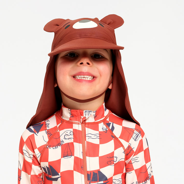 A smiling child wearing a brown bear-themed sun hat with ear flaps and a chin strap, paired with a red and white checkered zip-up swimsuit, against a plain light gray background.