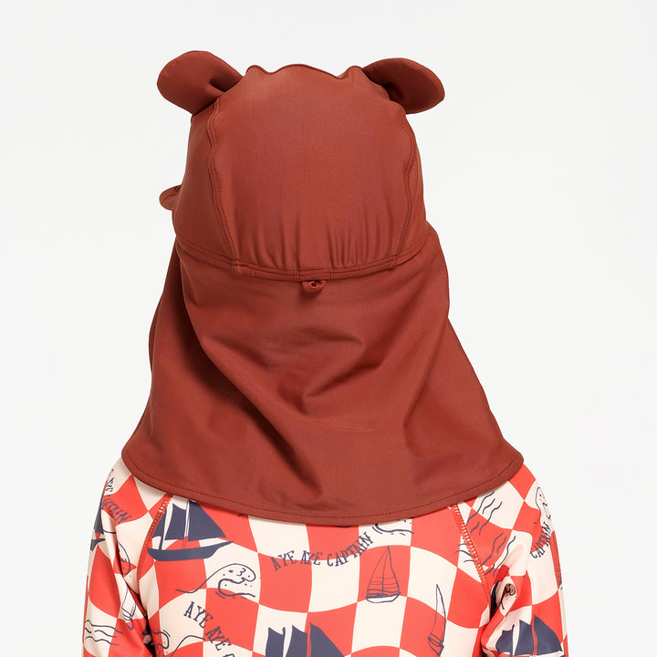 A child wearing a brown bear-ear sun hat and neck gaiter, paired with a red and white checkered swimsuit with nautical prints, shown from the back against a plain white background.